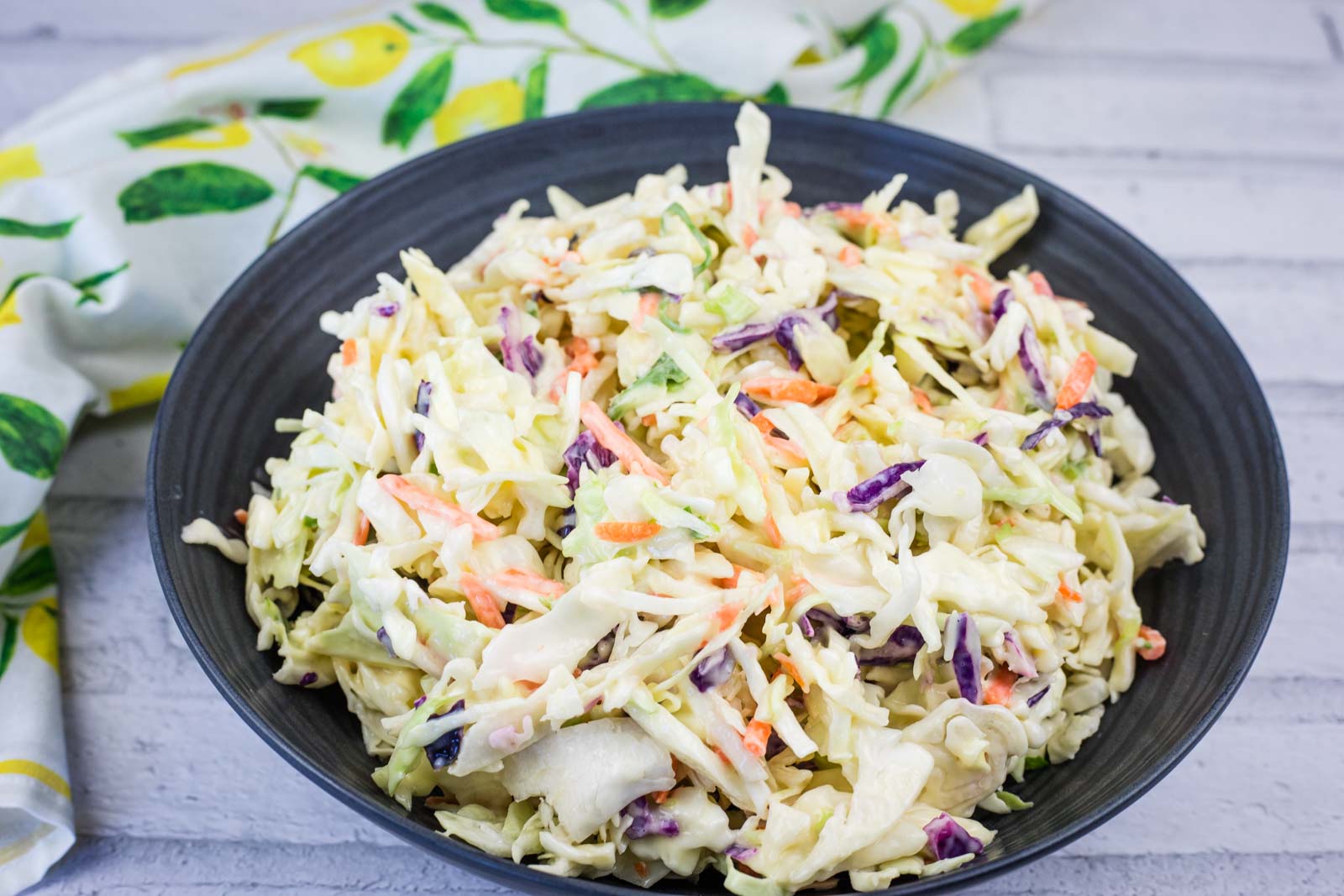 A black bowl filled with creamy coleslaw featuring shredded cabbage, carrots, and purple cabbage, placed on a light wooden surface with a colorful cloth nearby.