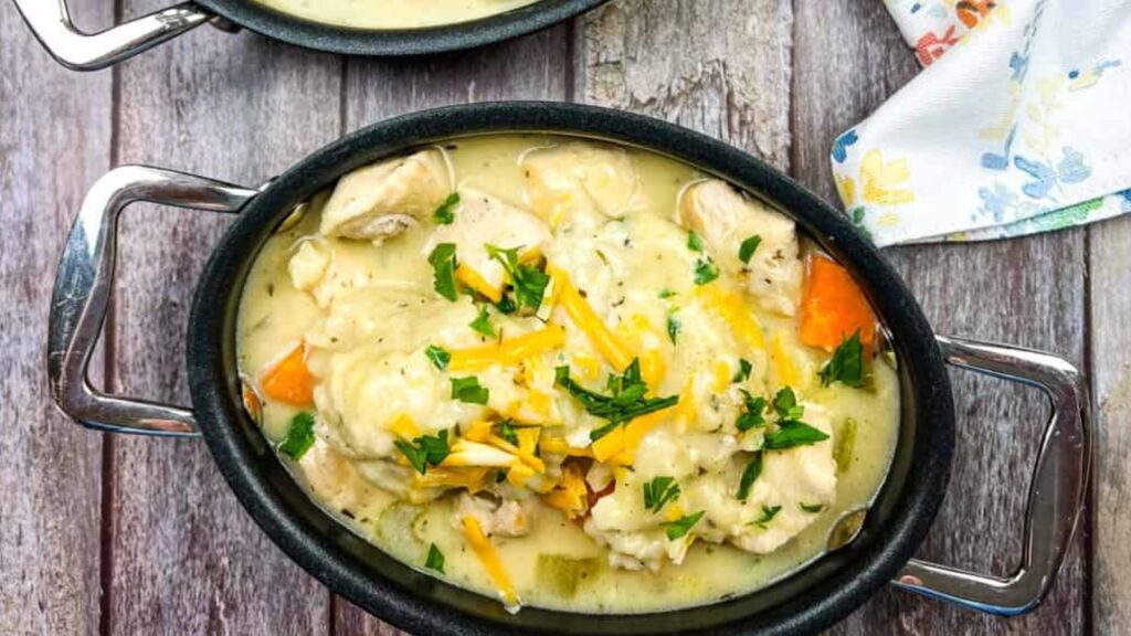 Oval dish containing chicken and dumplings in creamy sauce, topped with shredded cheese and chopped parsley, on a wooden table beside a floral napkin.